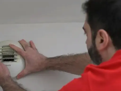 Un homme en chemise rouge effectue l'entretien d'une VMC murale, vérifiant le filtre encrassé.