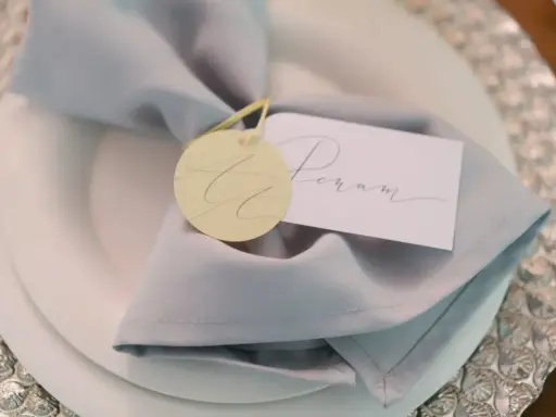 Elegancka winietka z imieniem "Peram" na jasnoszarym obrusie, ozdobiona okrągłym, żółtym tagiem.