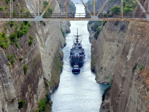 Una nave militare e un rimorchiatore navigano nel **Canale di Corinto**, tra alte pareti rocciose scavate dall'uomo.