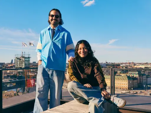 Två glada personer på en takterrass med utsikt över Stockholm, en solig dag. I bakgrunden syns stadens silhuett och en glimt av pelago slussen.