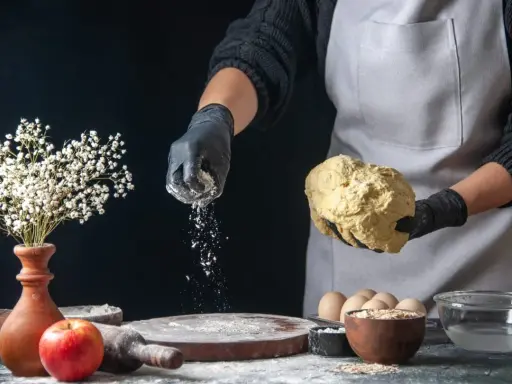Hände in schwarzen Handschuhen streuen selbsttreibendes Mehl über Teig. Ein Apfel und Blumen sind auf dem Tisch.