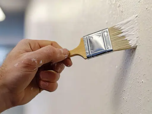 Mano aplicando pintura blanca con brocha, creando texturas. Descubre trucos para pintar paredes con efectos únicos.