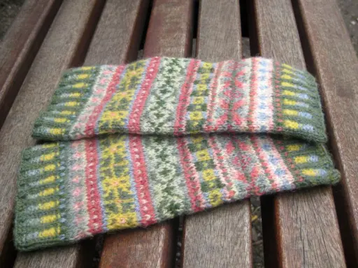 Två handledsvärmare i fair isle stickning med mönster i grönt, rosa, gult och blått ligger på en träbänk.