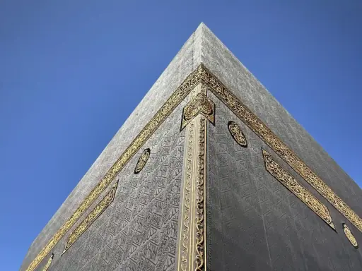 Die Kaaba in Mekka, Saudi-Arabien, mit ihrer schwarzen Hülle und goldenen Verzierungen vor blauem Himmel.
