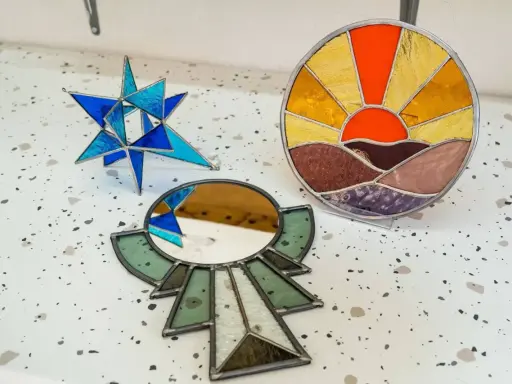 Trois pièces d'art en vitrail : une étoile bleue, un soleil couchant et un miroir ornemental. Créations d'un atelier vitrail Paris.