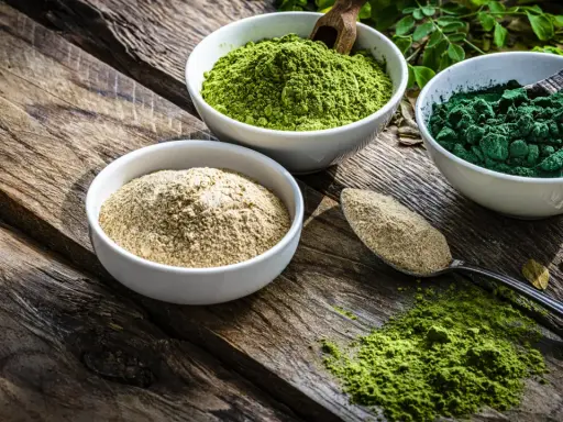 Drei Schalen mit Pulver auf Holz: hellbraun, leuchtend grün und dunkelgrün. Spirulina gesund und voller Nährstoffe.