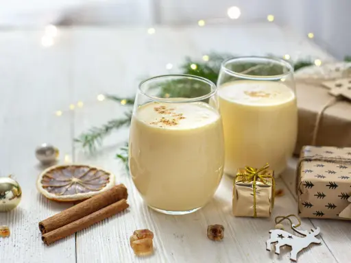 Zwei Gläser cremiger Cocktail mit Eigelb, garniert mit Zimt, umgeben von Weihnachtsdekorationen und Geschenken.