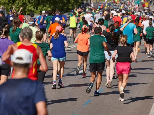 Tłum biegaczy pokonuje kolejne kilometry maratonu. Widać ich nogi w ruchu, kolorowe stroje i cienie na asfalcie.