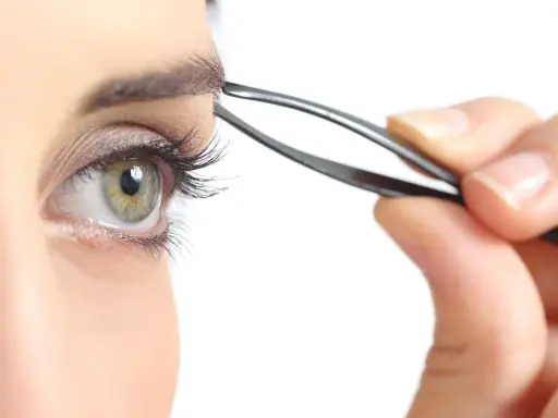 Une main utilise une pince à épiler pour parfaire l'épilation des sourcils d'un œil vert aux longs cils.
