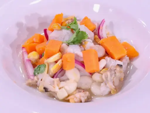 Delicioso ceviche de corvina con trozos de camote, choclo, cebolla roja y culantro, servido en un plato blanco.