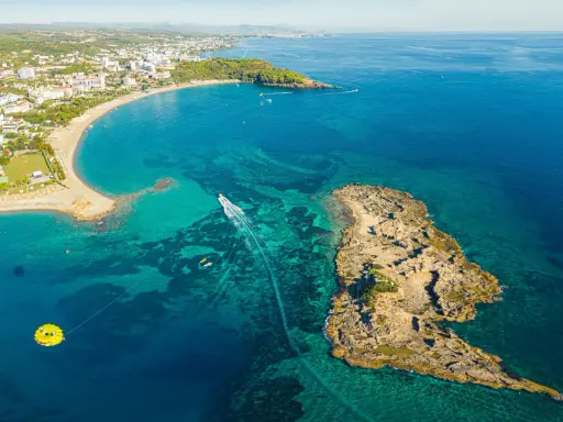 Widok z lotu ptaka na wybrzeże z hotelami, plażą i turkusową wodą. Widać łódź, parasailing i skaliste wysepki, które są świetnymi okurcalar atrakcje.