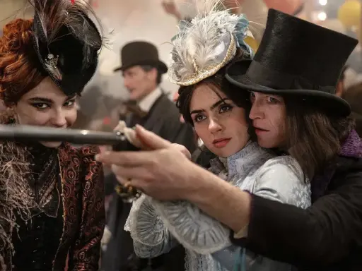 Una donna con un cappello piumato punta una pistola, protetta da un uomo con un cilindro. Un'altra donna con un cappello decorato sorride. Un momento intenso da "Dracula a love tale".