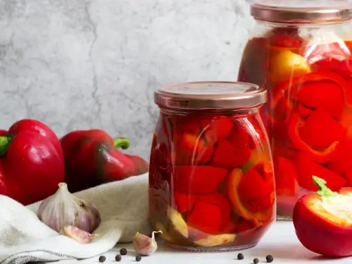 Zwei Gläser mit eingelegter Paprika, daneben frische rote Paprika, Knoblauch und Pfefferkörner. Perfekt zum Paprika einkochen.
