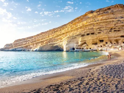 Ein malerischer Strand auf Kreta mit türkisfarbenem Wasser, das sanft an den Kiesstrand schwappt. Die beeindruckende Felswand mit ihren Höhlen bietet eine einzigartige Kulisse.