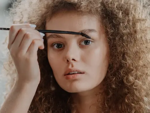 Une jeune femme aux cheveux bouclés applique un produit sur ses sourcils décolorés avec une brosse.