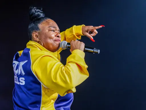 Roxanne Shanté w żółtej kurtce z niebieskimi wstawkami śpiewa do mikrofonu, wskazując palcem.