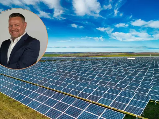Uśmiechnięty mężczyzna w garniturze na tle farmy fotowoltaicznej. Obraz symbolizuje produkcję energii z fotowoltaiki w poszczególnych miesiącach.