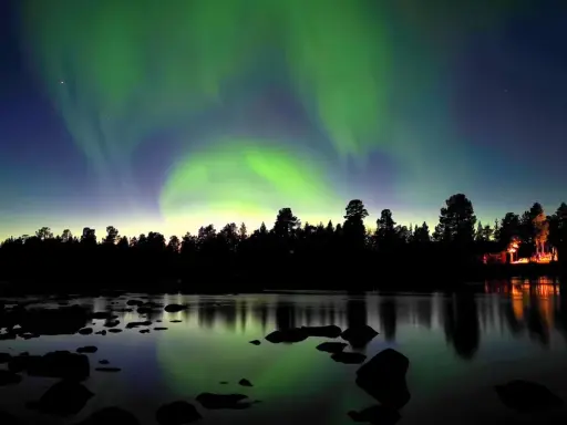 Norrsken dansar över en vinterlandskap vid Kiruna. En skogskant och en sjö med stenar speglar himlens magiska ljus.