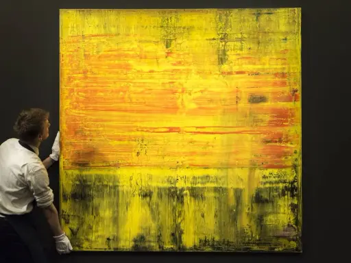 A man in white gloves carefully handles a large abstract painting with vibrant yellow and orange hues, a testament to Gerhard Richter's net worth.