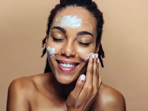 Une femme applique de la crème sur son visage. Elle sourit, les yeux fermés. Elle se demande que veut dire indice 50 pour sa protection solaire.
