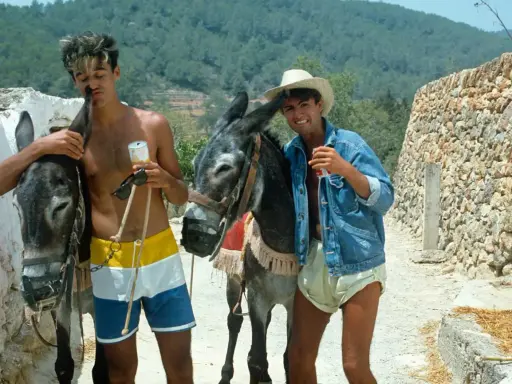 Tony Pike Ibiza: due ragazzi con asini, uno con pantaloncini colorati, l'altro con giacca di jeans e cappello di paglia, sorseggiano bevande.