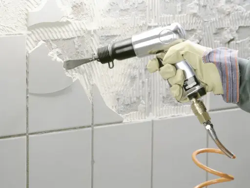 Mit einem Pressluftmeißel werden weiße Fliesen entfernt. Die Wand darunter ist bereits freigelegt und zeigt Mörtelreste.