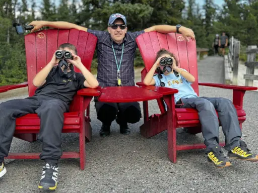 Un padre y sus dos hijos disfrutan de la naturaleza en Canadá con niños, observando el paisaje con binoculares.