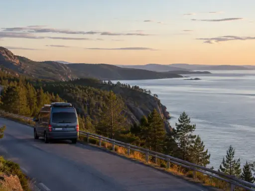 En husbil kör längs en slingrande väg vid den vackra Höga kusten. Solnedgången färgar himlen i mjuka toner över det glittrande havet.