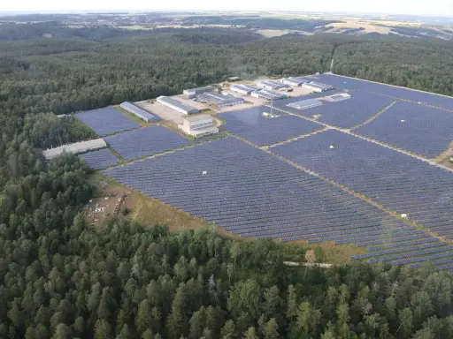 Große Solaranlage im Wald, die Sonnenenergie sammelt. Gebäude und Reihen von Solarmodulen erstrecken sich über die Landschaft.