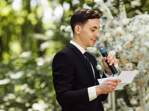 El maestro de ceremonias, con traje negro y micrófono, lee un discurso frente a un arco de flores.