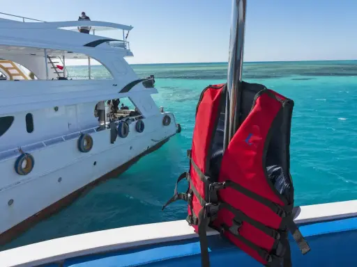 Gilet de sauvetage rouge prêt pour l'aventure en mer, à côté d'un yacht blanc. Les **œuvres mortes du bateau** se reflètent dans l'eau turquoise.