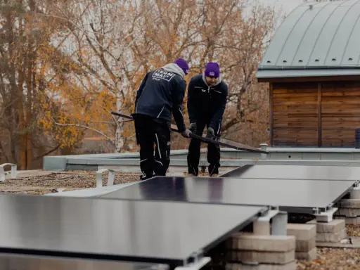 Två personer installerar solceller på ett papptak. Höstfärgade träd syns i bakgrunden.