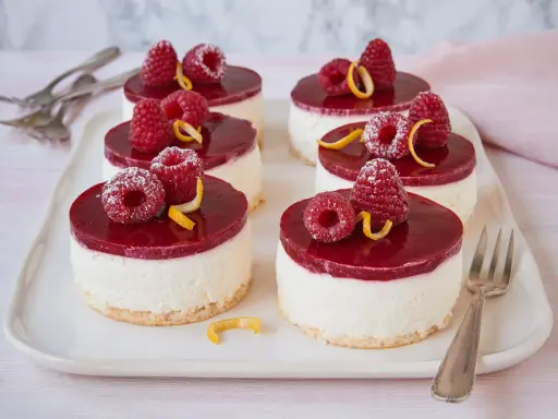 Sechs kleine Kuchen mit Himbeeren und Zitronenzesten auf einem weißen Teller.