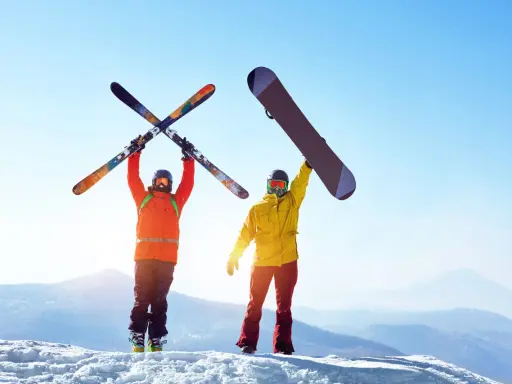 Dwóch narciarzy na szczycie góry, jeden z nartami, drugi z deską snowboardową, cieszy się słońcem i widokiem.