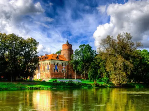 Malowniczy zamek w Łódzkiem nad rzeką, otoczony zielenią i chmurami.
