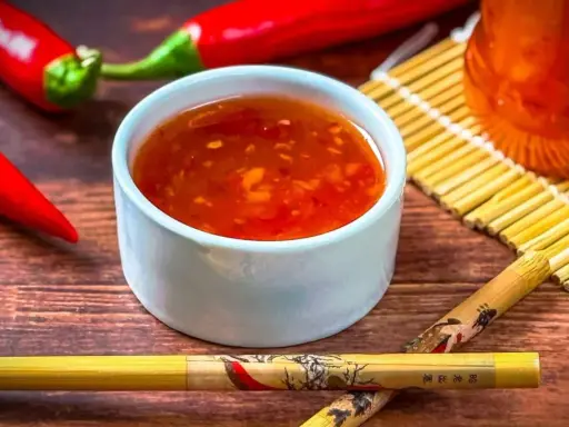 Schüssel mit leuchtend roter Sweet Chilli Sauce, daneben rote Chilis und Stäbchen. Perfekt für dein Sweet Chilli Sauce Rezept!