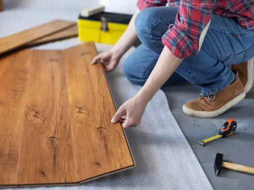 Persona instala tarima flotante, uniendo tablas de madera con cuidado. Herramientas como martillo y cinta métrica a mano.