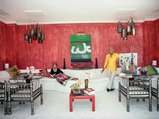 Interni di case anni '70: una donna legge sul divano bianco, un uomo mostra un quadro. Pareti rosse, lampade a grappolo.