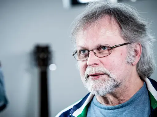 Clark Olofsson idag, med grått hår, skägg och glasögon, blickar mot betraktaren.