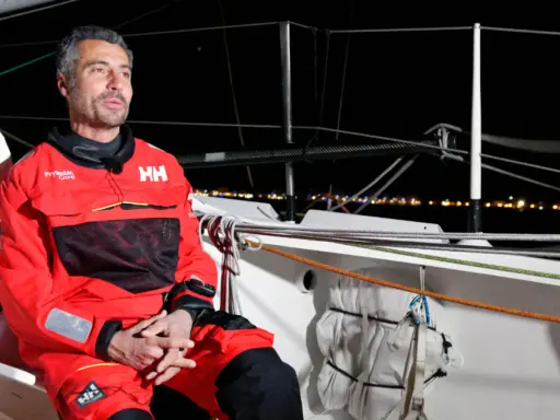 Giancarlo Pedote, skipper en tenue de voile rouge, sur un bateau la nuit, avec des lumières de ville en arrière-plan.