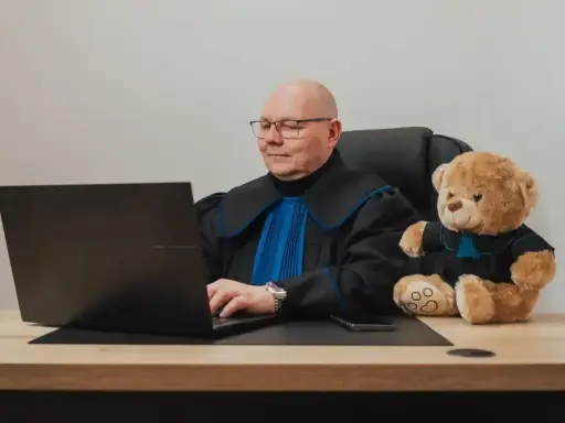 Prawnik w todze z pluszowym misiem obok, pracujący przy laptopie. Zastanawiasz się, ile kosztuje porada prawna?