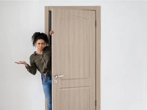 Mujer asomándose por una puerta de madera clara, con expresión de duda. Los colores de puertas como esta son muy versátiles.