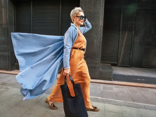 Elegancka kobieta w pomarańczowym garniturze i błękitnym płaszczu z wiatrem we włosach. Inspiracja dla mody dla starszych panów.