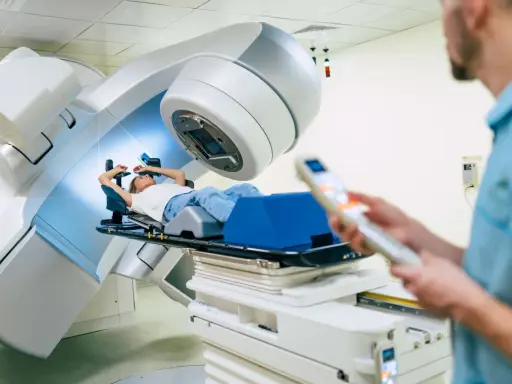 Ragazza sdraiata su lettino per radioterapia, tecnico in primo piano controlla il macchinario. Speranza di guarigione, ma con il timore dei radioterapia danni permanenti.