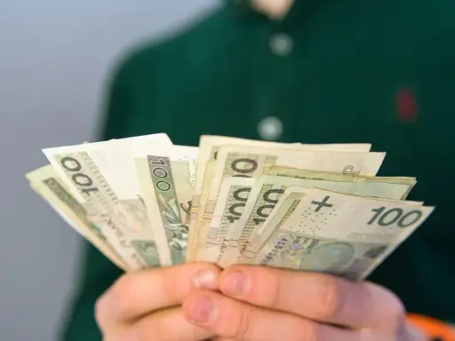 Dłonie trzymają wachlarz banknotów 100 zł. Może to być pomoc finansowa, np. renta po zmarłym ojcu, która zapewnia wsparcie.