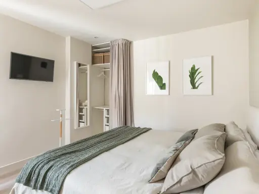 Ein offener Kleiderschrank, der wie ein Kleiderschrank selber bauen mit Vorhang wirkt, mit Regalen und Kleiderstange. Daneben ein Spiegel und ein Bett mit grüner Decke.