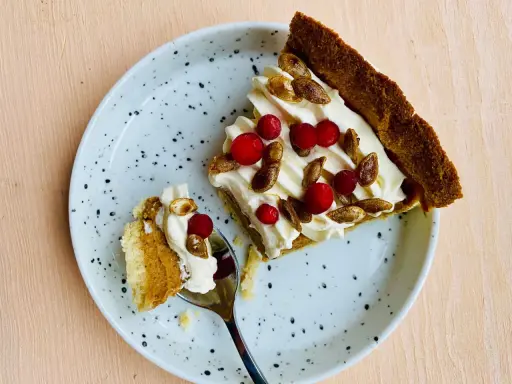 En bit glutenfri pumpapaj med vispgrädde, lingon och rostade pumpafrön på en prickig tallrik. En sked med en bit paj ligger bredvid.