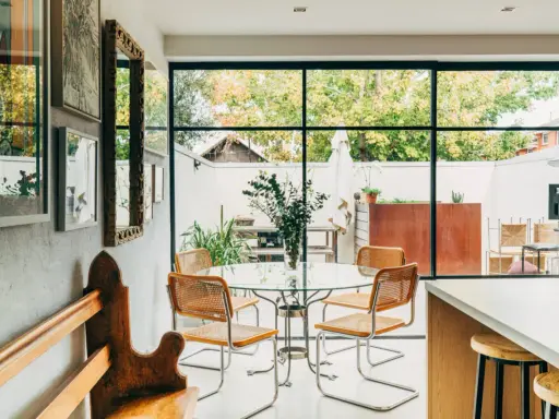 Sedie in paglia e metallo, ispirate a Marcel Breuer, attorno a un tavolo di vetro in una luminosa sala da pranzo con vista sul giardino.