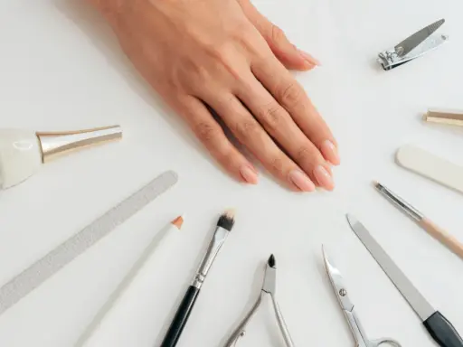 Dłoń z naturalnymi paznokciami otoczona akcesoriami do manicure. Wśród nich pilnik do paznokci naturalnych, cążki, nożyczki i pędzelki.