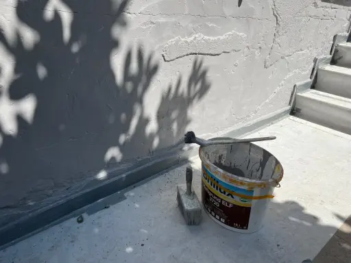 Werkzeug und Farbeimer bereit zum Ausbessern des Sockelputzes. Schatten von Blättern fallen auf die Wand.
